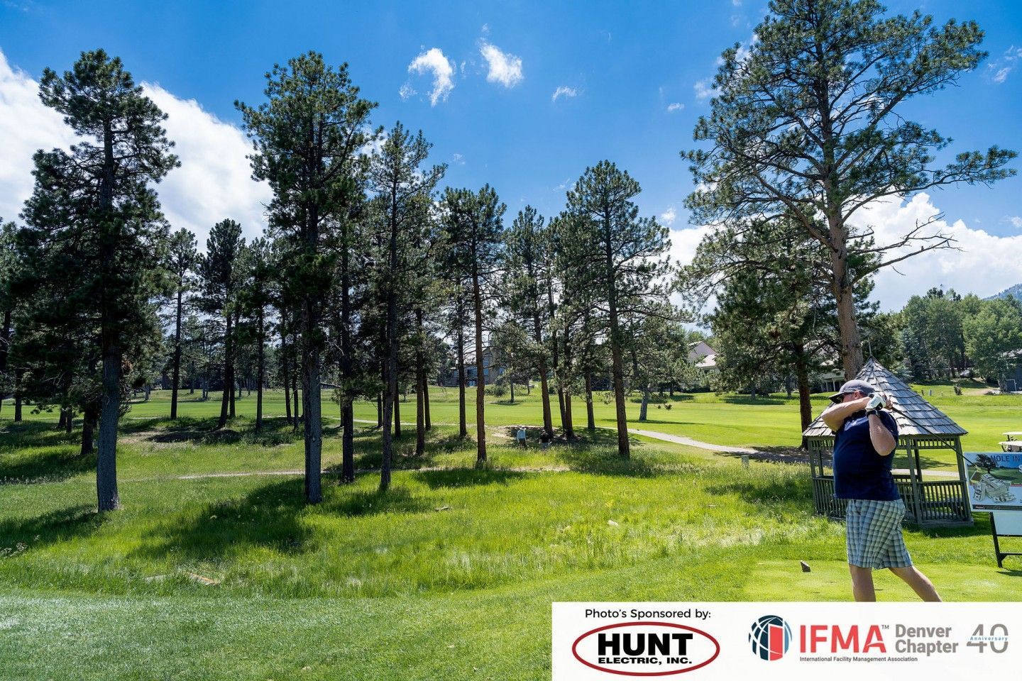 Golfer tees off on a sunny golf course, surrounded by trees and a gazebo.