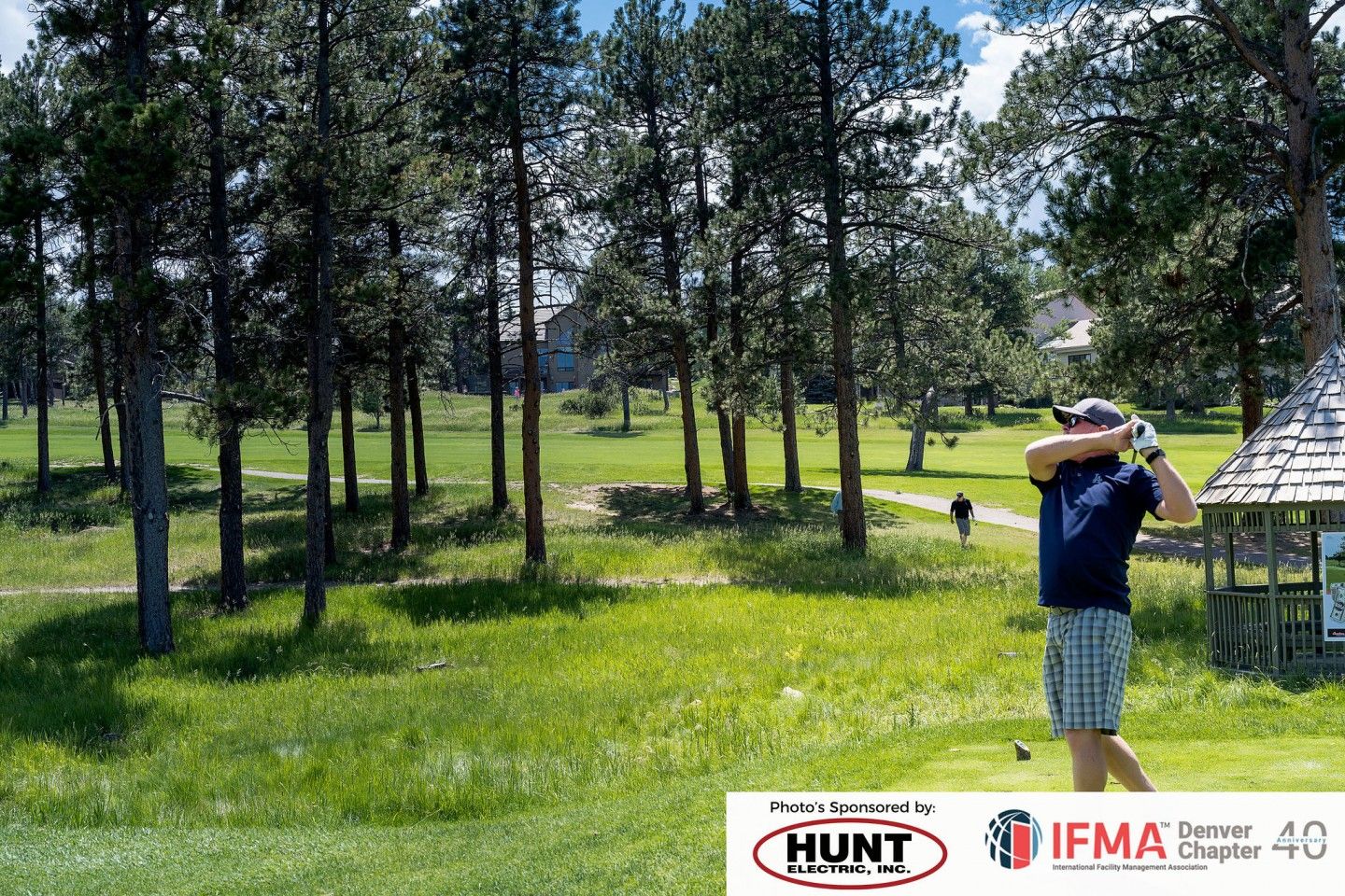 Golfer swings on a green golf course, surrounded by trees.