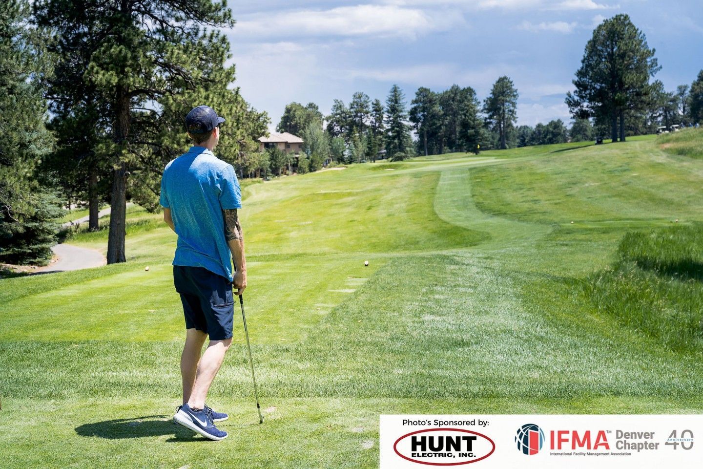 Man on a golf course holding a club, looking toward the green. Sunny day, trees, and fairways visible.