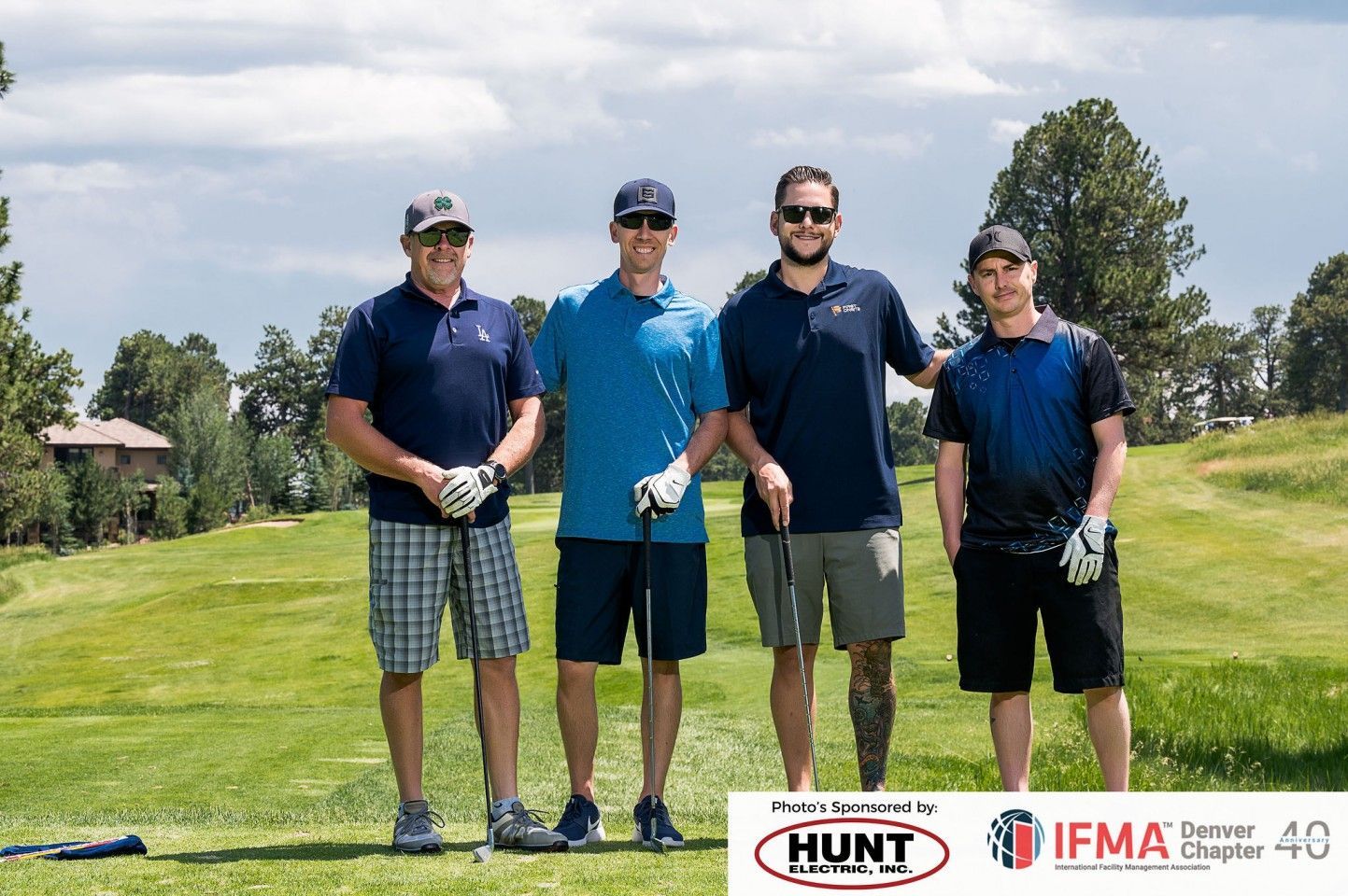 Four men on a golf course pose for a photo. They are wearing golf attire, holding clubs. Sunny day.