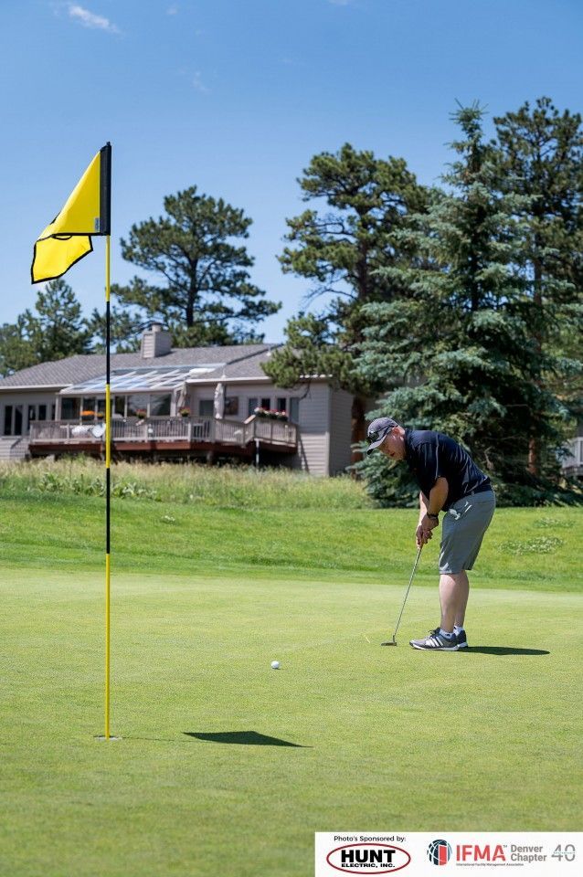 Man putting a golf ball on a green, with flag, house and trees visible under a blue sky.
