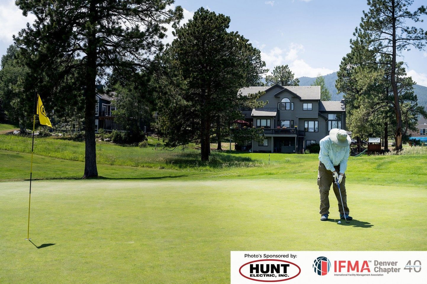 Golfer putting on green, flag in the distance, trees and a house in background.