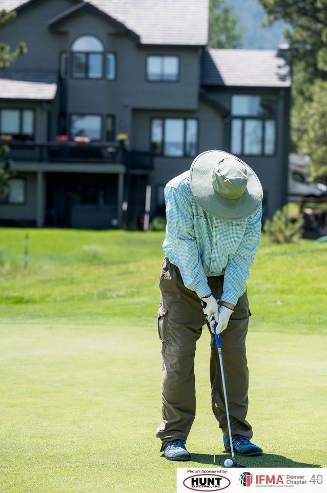 Man in a hat playing golf on a green course; large gray house in the background.