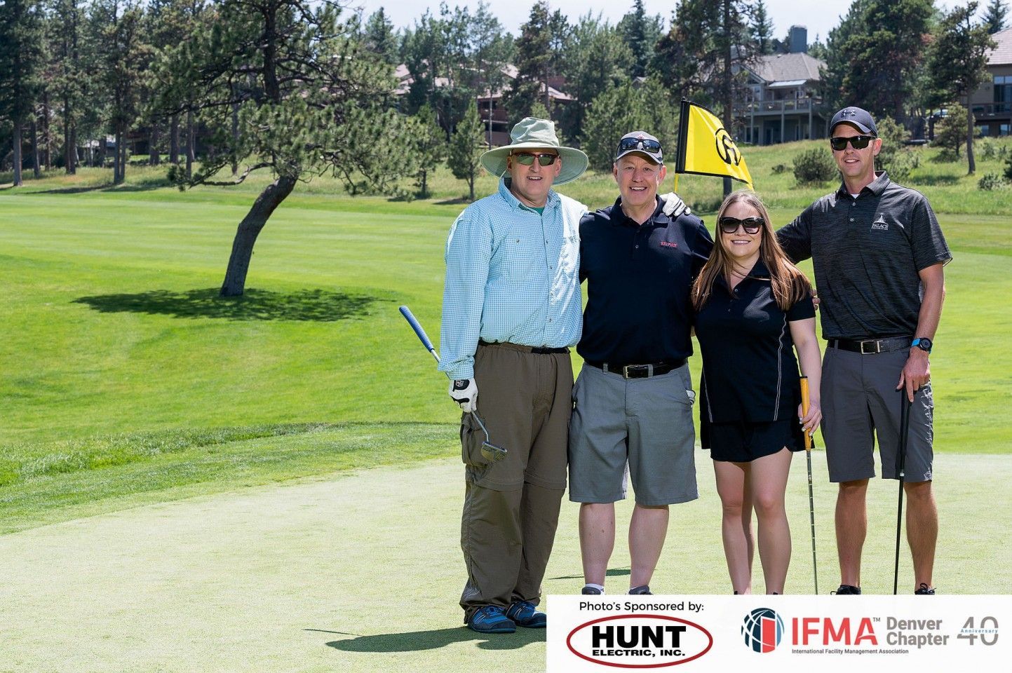 Four people pose on a golf course, one woman, two men in shorts, one man in a hat. A green flag in the background.