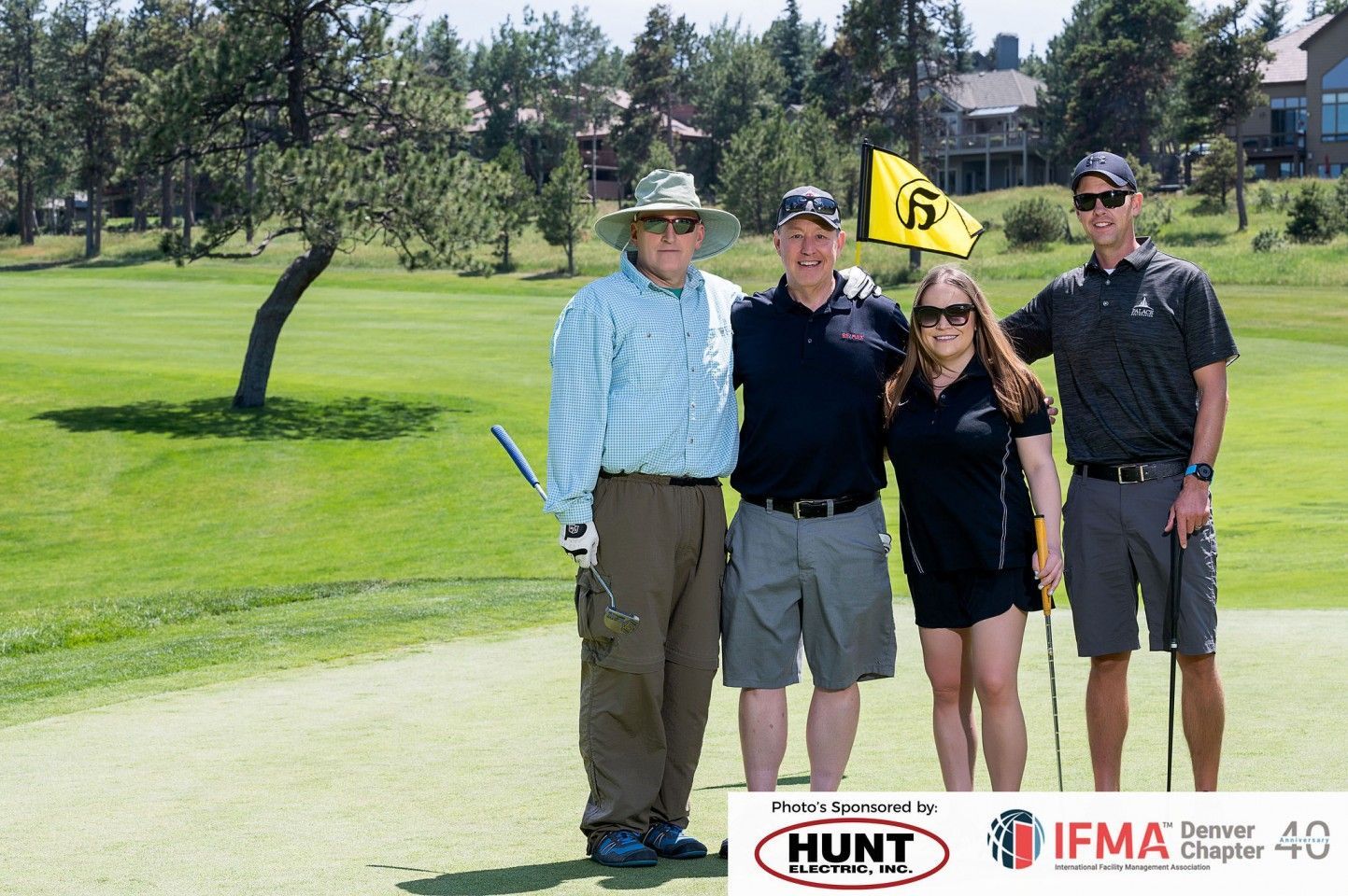 Four people pose on a golf course: a man in a hat holds a club, while three others smile.