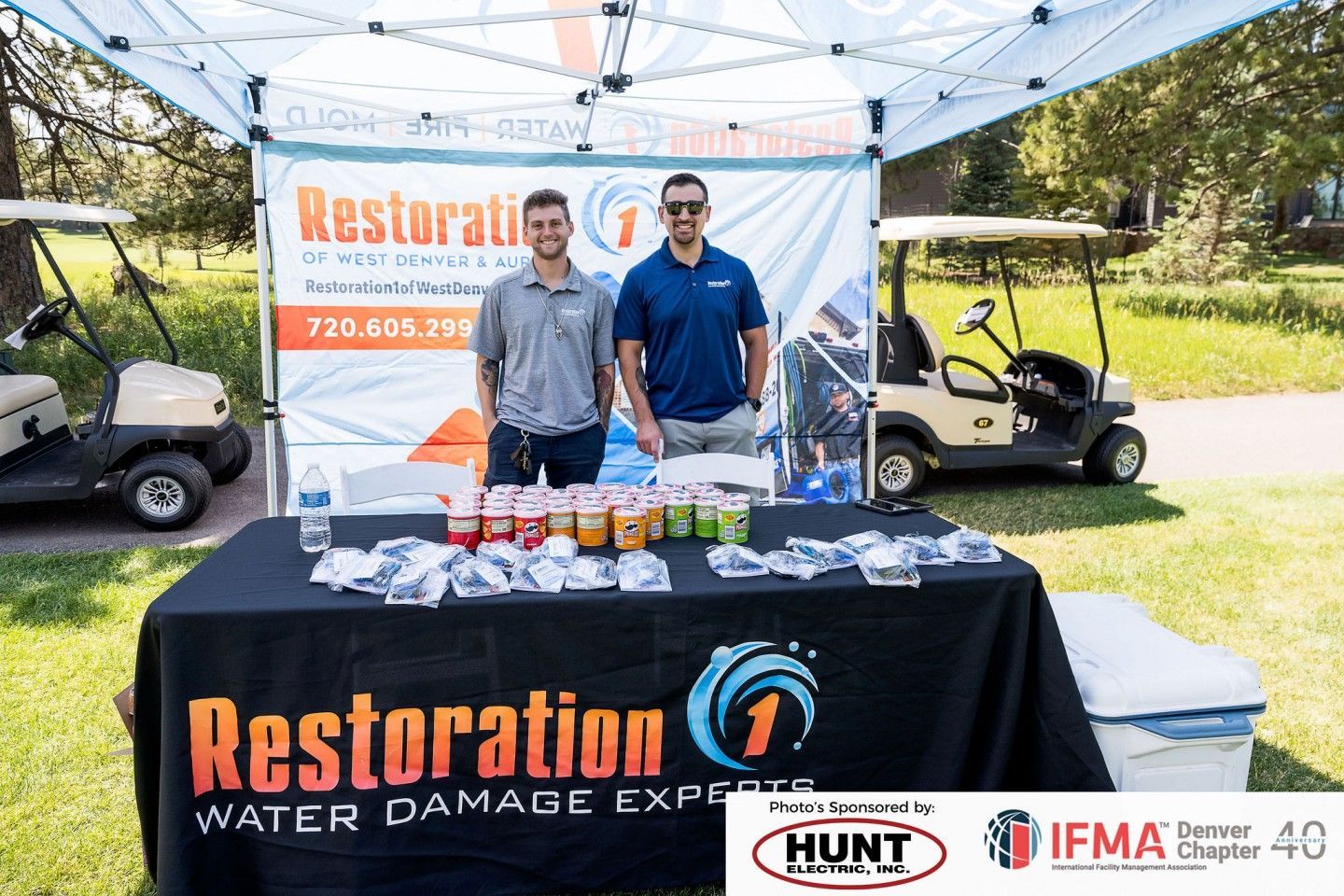 Two men stand behind a booth for Restoration 1, a water damage expert, at an outdoor event.