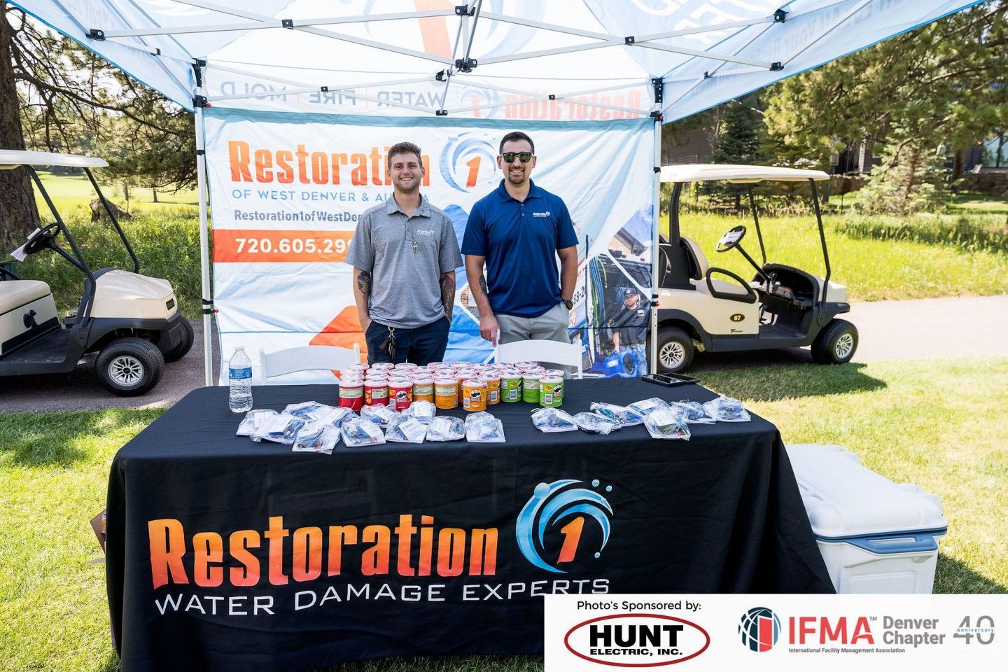 Two men at a Restoration 1 booth at an outdoor event with drinks, golf carts, and logo signage.