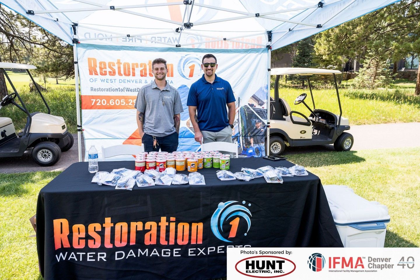 Two men at a Restoration 1 booth at an outdoor event, serving drinks next to a golf cart.