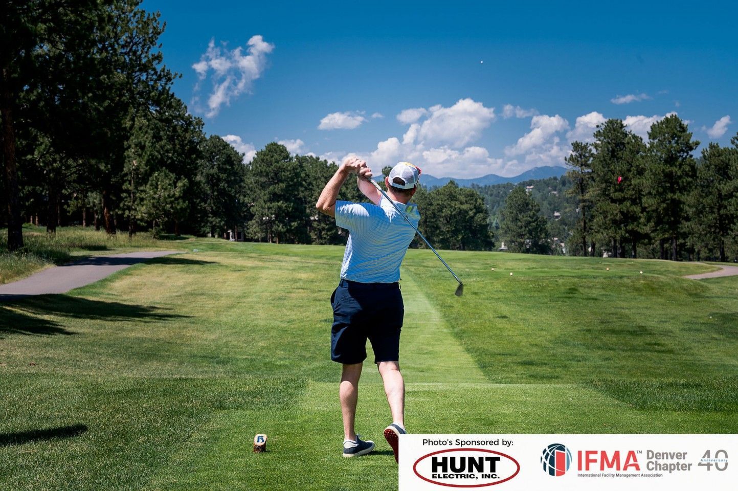 Golfer swings club on a sunny green, trees and mountains in background.
