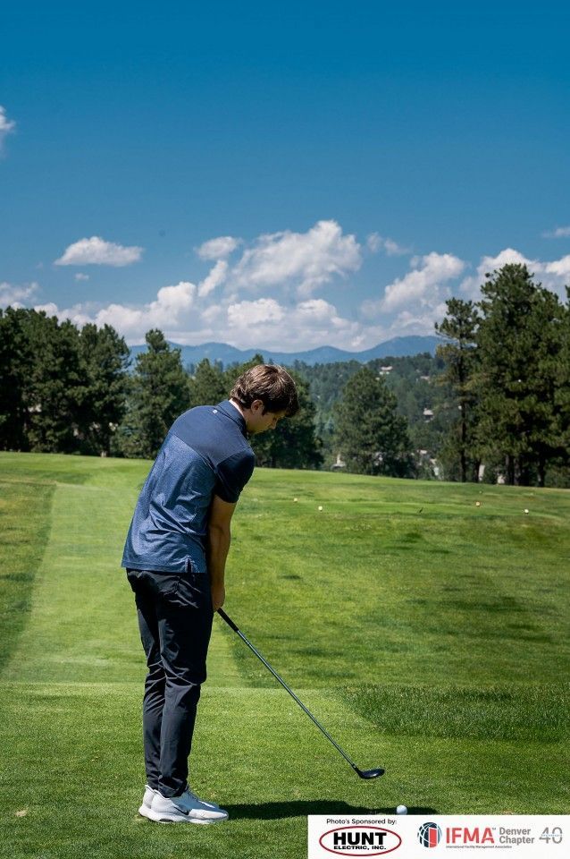 Golfer swings club on green; blue sky, trees, and mountains in the background.
