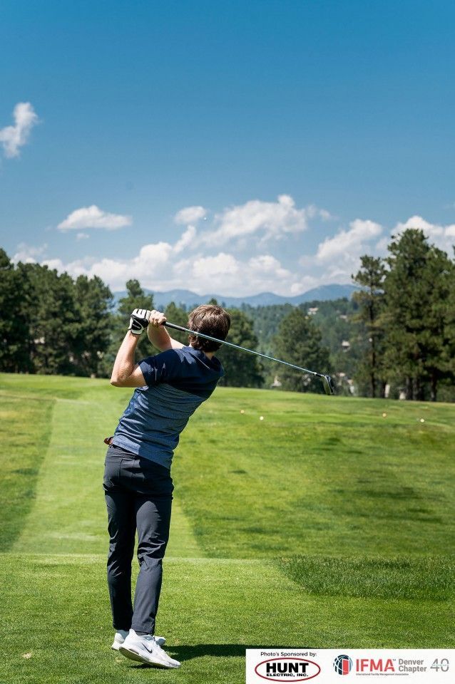 Golfer swinging club on a sunny golf course. Green grass, blue sky, trees, and mountains in background.