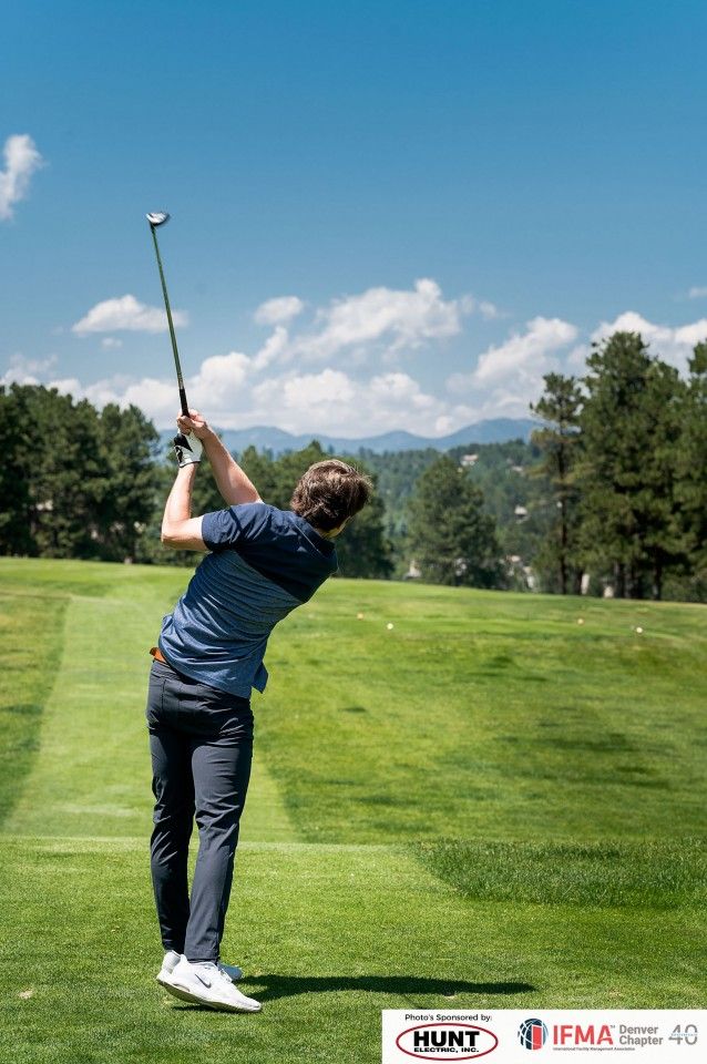Golfer swinging a club on a green course, with blue sky and trees in the background.