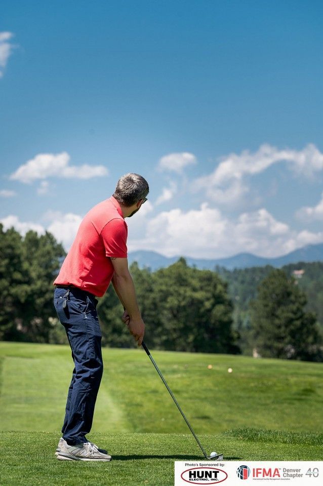 Man in red shirt preparing to swing golf club on green course under a blue sky.