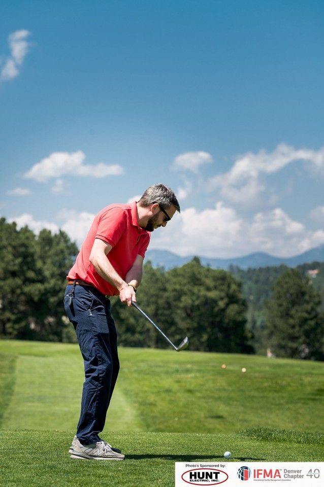 Man in red shirt swings golf club on green course under a blue sky.