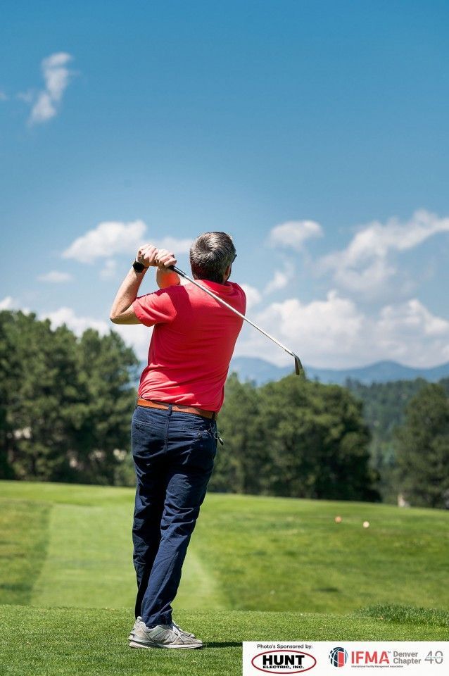 Golfer in red shirt swings a club on a green course, blue sky and trees in the background.