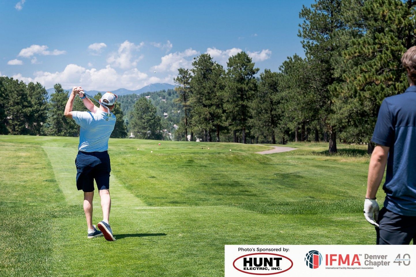 Golfer swings club on green course with trees, another person watches. Blue sky, mountains.