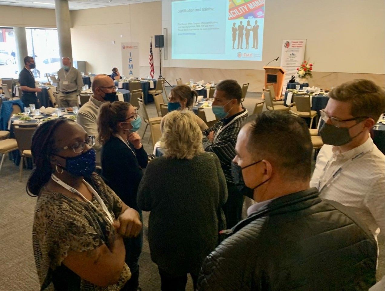 Group of people in masks talking at a conference, some standing and others sitting at tables.