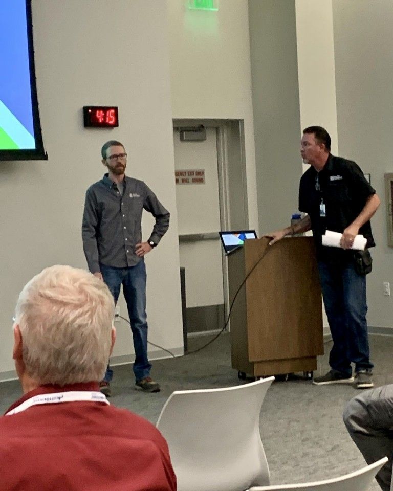 Two men at a podium giving a presentation. One man stands with his hands on his hips, and the other man holds papers.