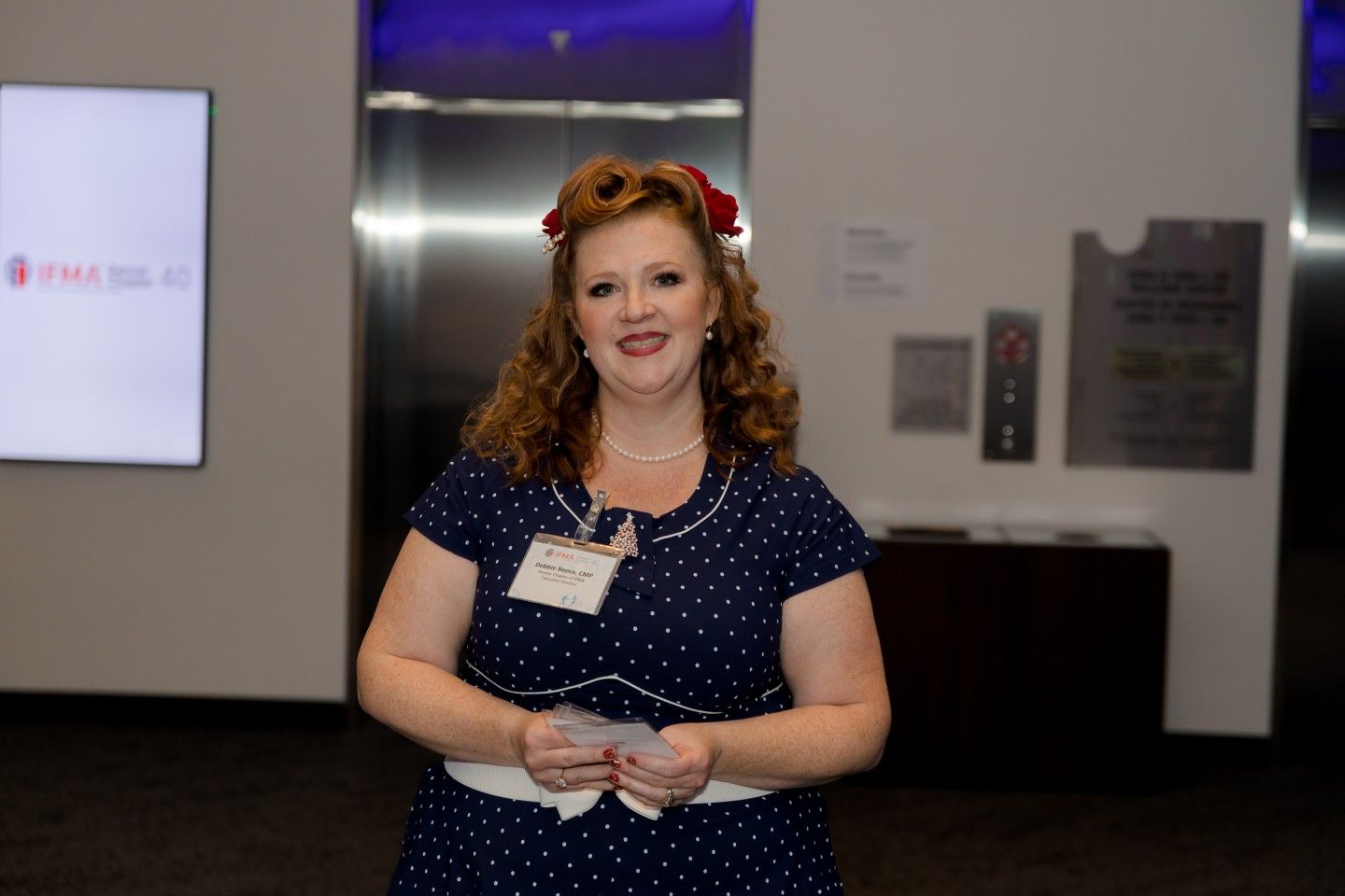 Woman with red hair in a blue polka-dot dress, smiling, holding cards, in front of elevators.