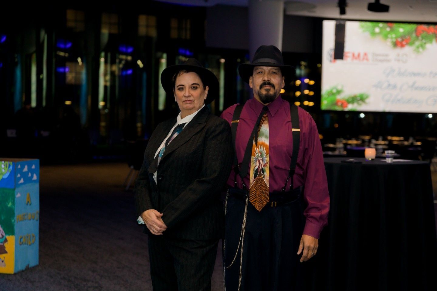 Two people in fedoras and formal attire stand at an event; a welcome banner is visible in the background.