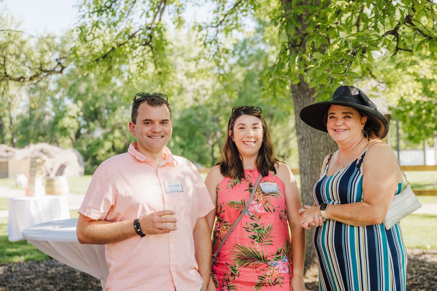 Three people smiling outdoors near a table. Man in pink shirt, woman in floral dress, woman in striped dress and hat.