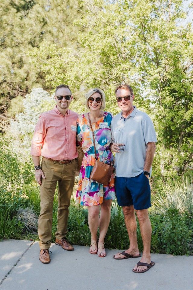 Three people smiling outdoors: woman in colorful dress, men in casual attire.