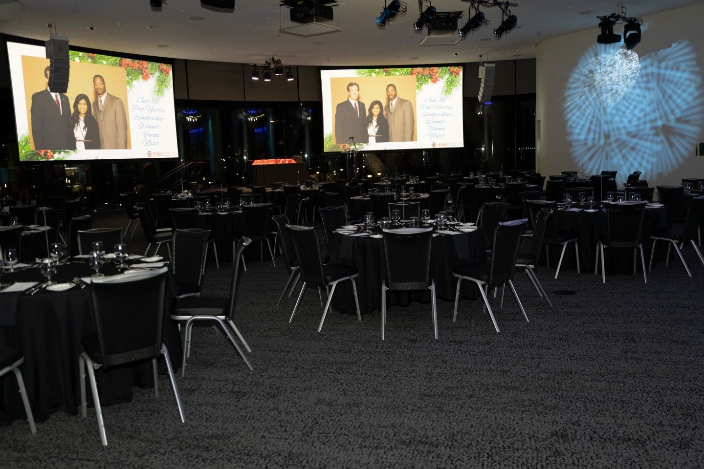 Banquet hall with round tables, black linens, chairs, and two screens displaying photos.