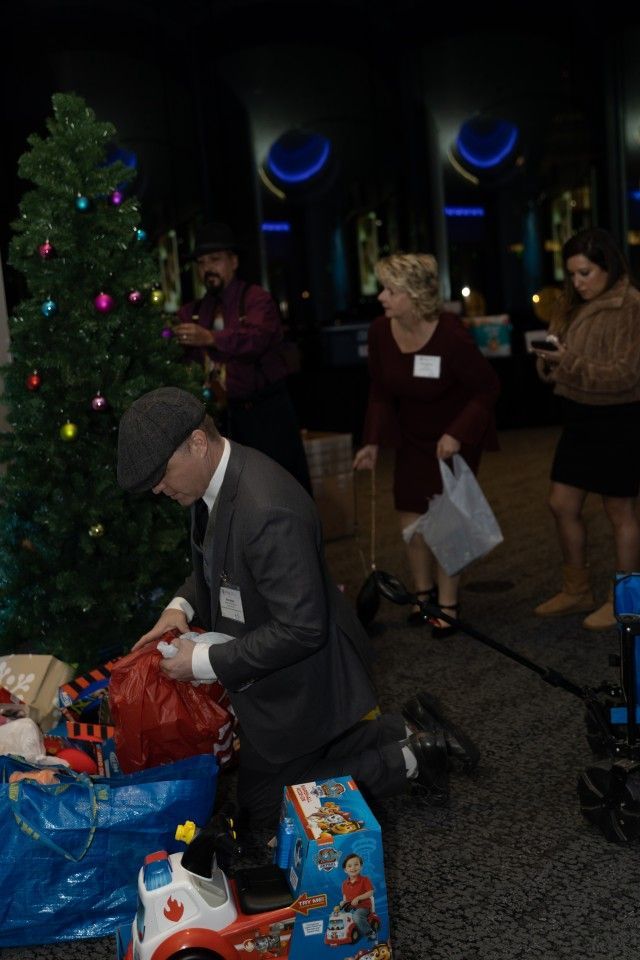 Man in suit kneeling by a Christmas tree, sorting toys. Others in the background watch.