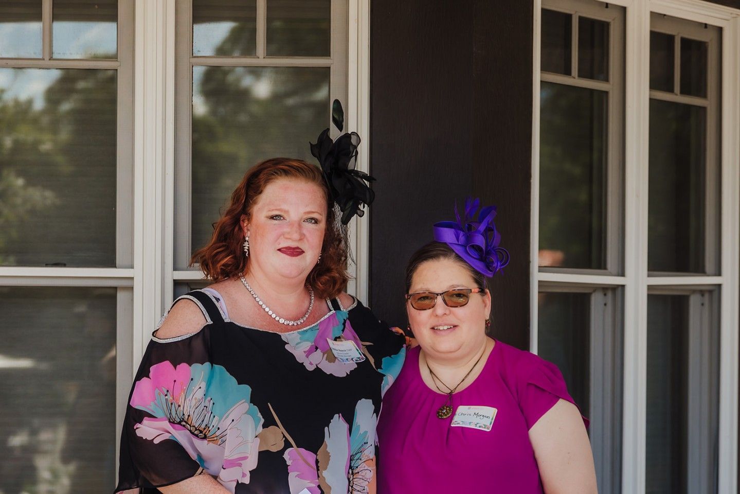 Two women smiling, posing in front of a window. One in a floral top, the other in purple with a hat.