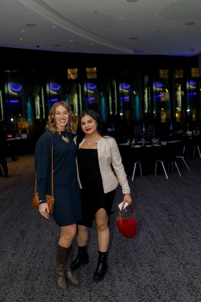 Two women posing indoors, one in blue dress and boots, the other in black dress and jacket, smiling.