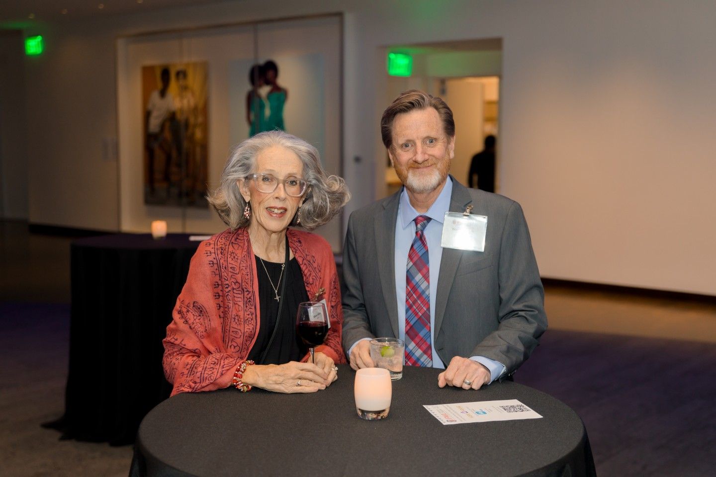 Two people smile at a table with drinks in a room. A woman wears a red shawl. Man in a suit with a striped tie.