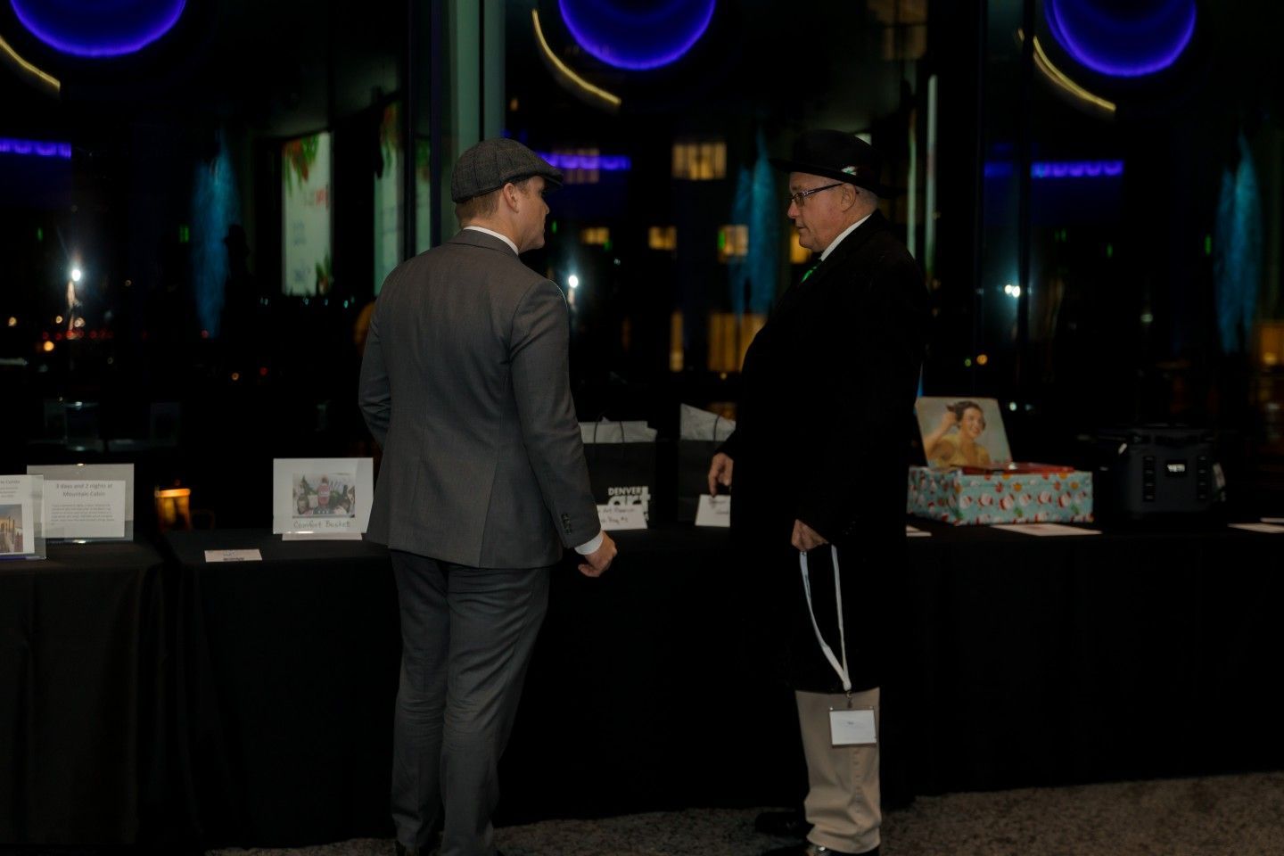 Two men in suits converse in front of a table with items, likely at an event with dark background.