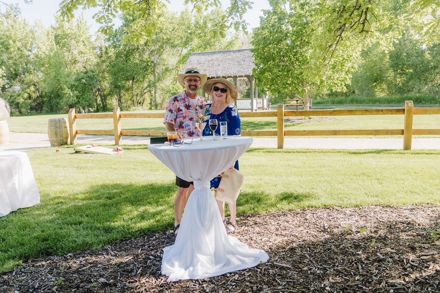 Couple stands behind a small table outdoors, smiling. The man wears a Hawaiian shirt, the woman a blue top.