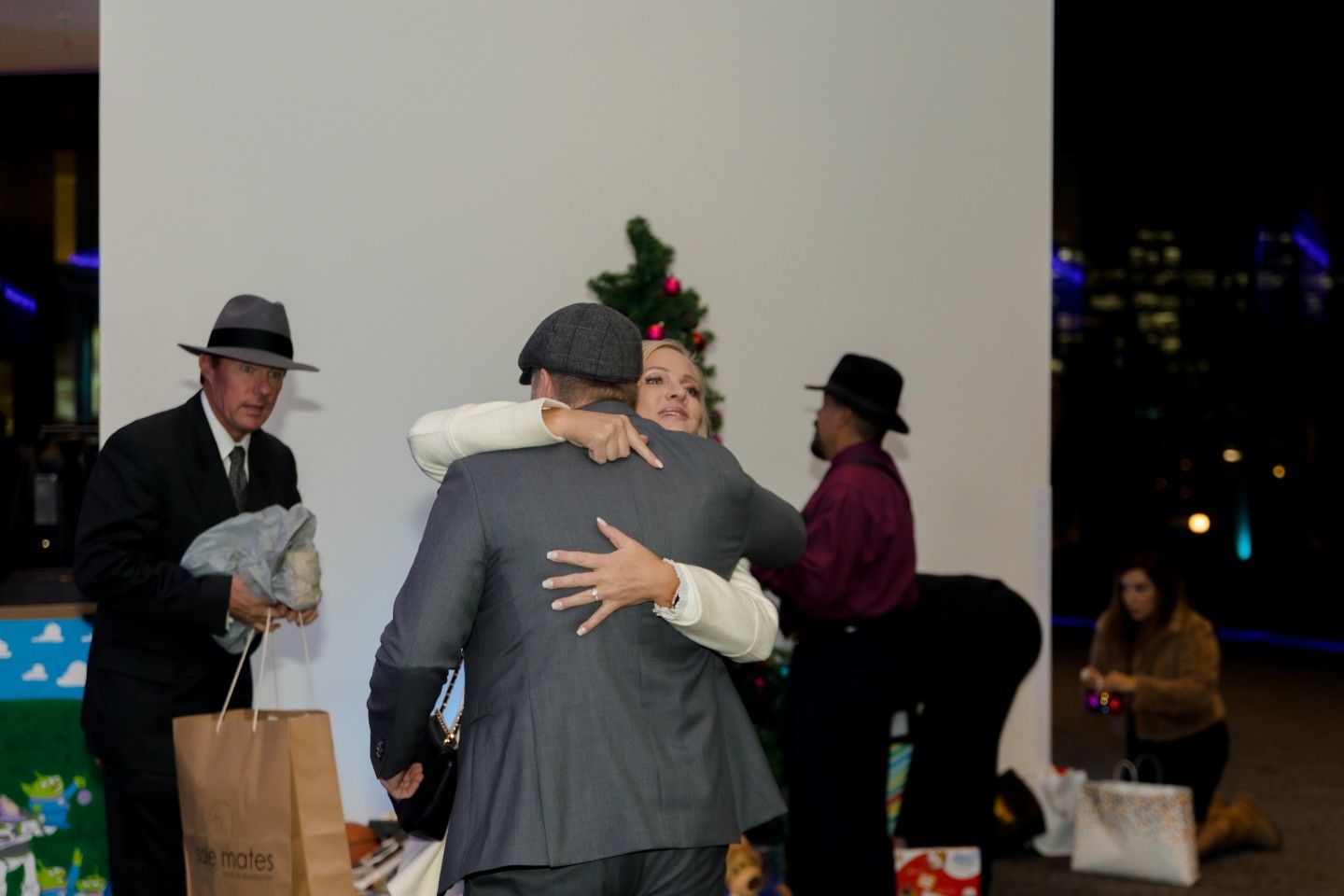 People hugging by a small Christmas tree, some wearing hats, others holding gifts, night setting.