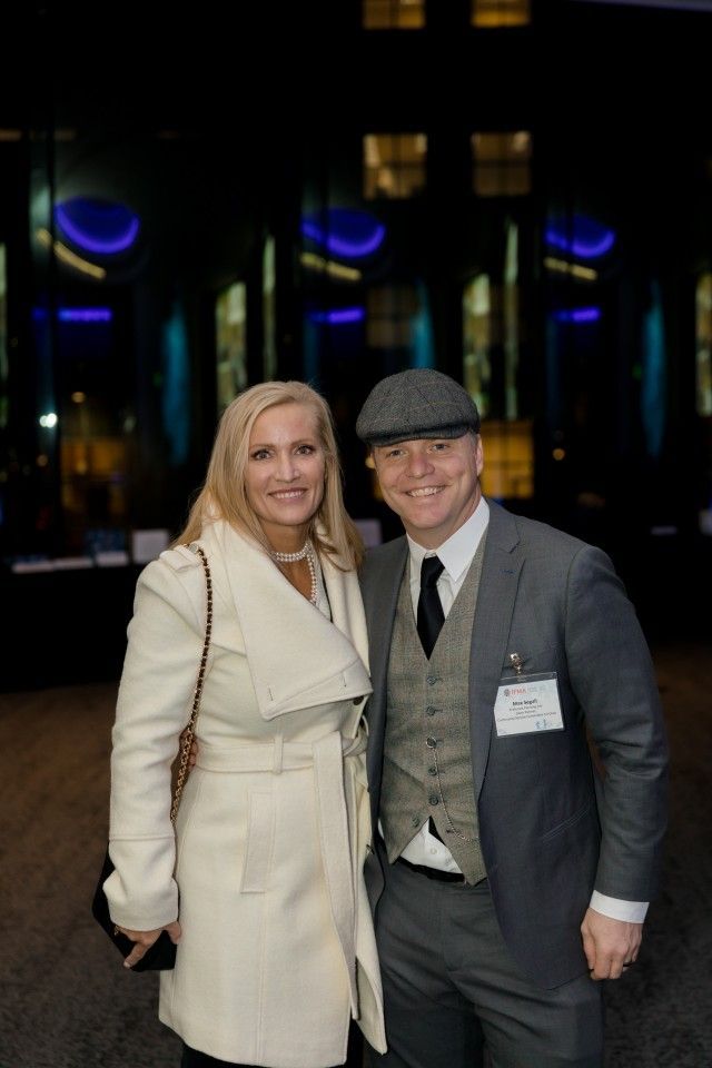 Woman in cream coat and man in gray suit pose together at an event.