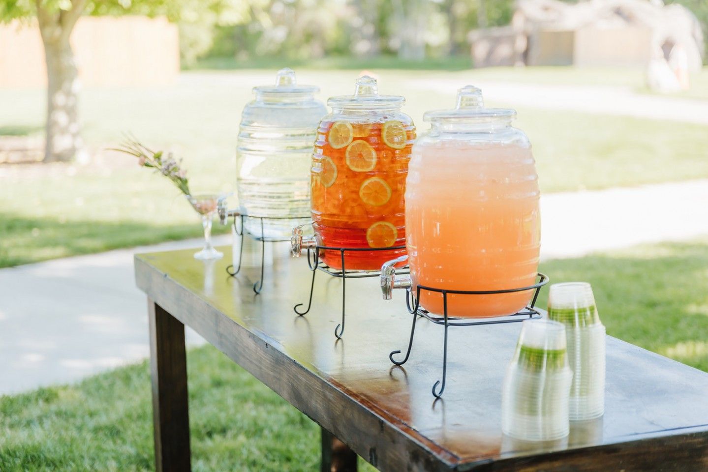 Three glass drink dispensers with different colored beverages on a wooden table outdoors.