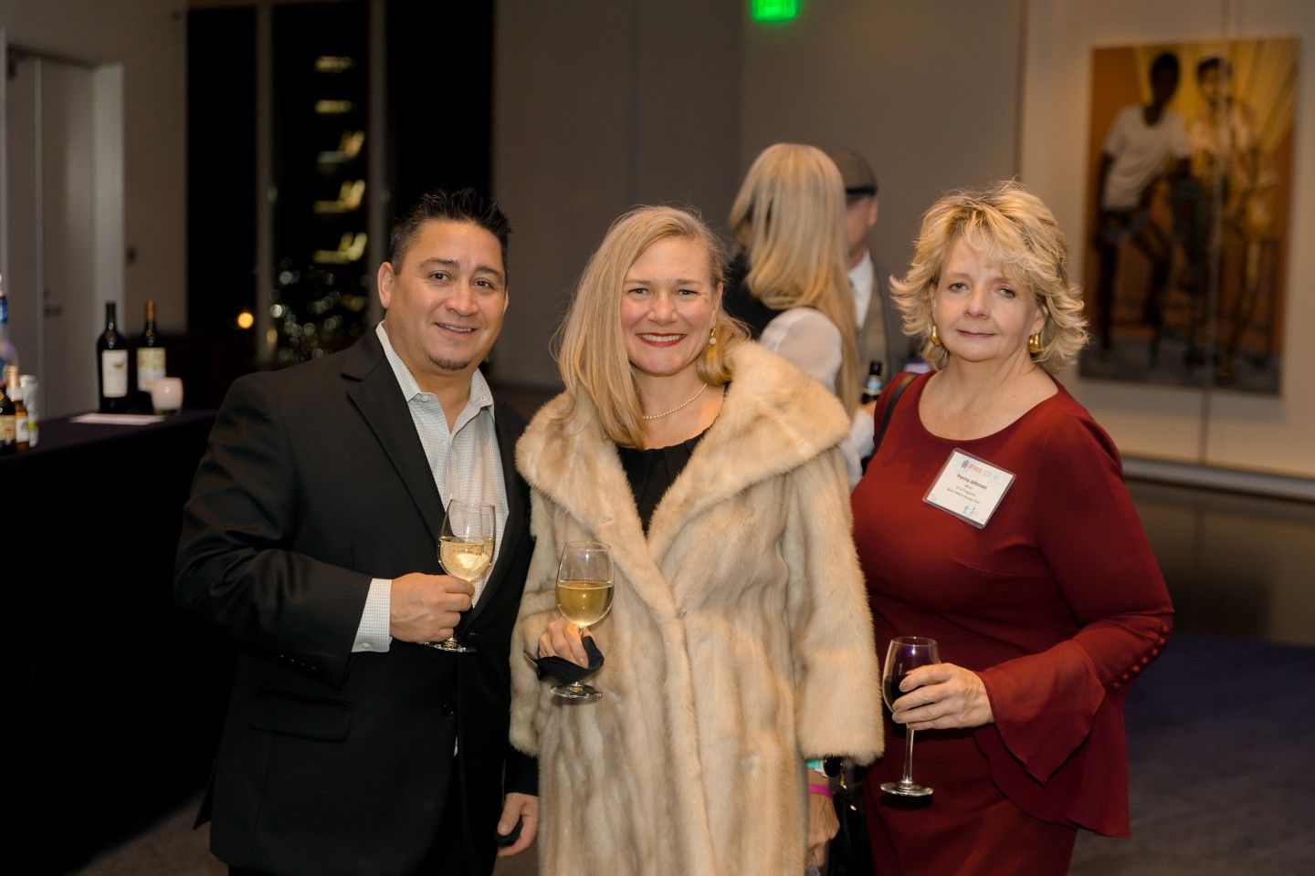 Three people smiling, holding drinks, at a formal event. Woman in fur coat, man in suit.