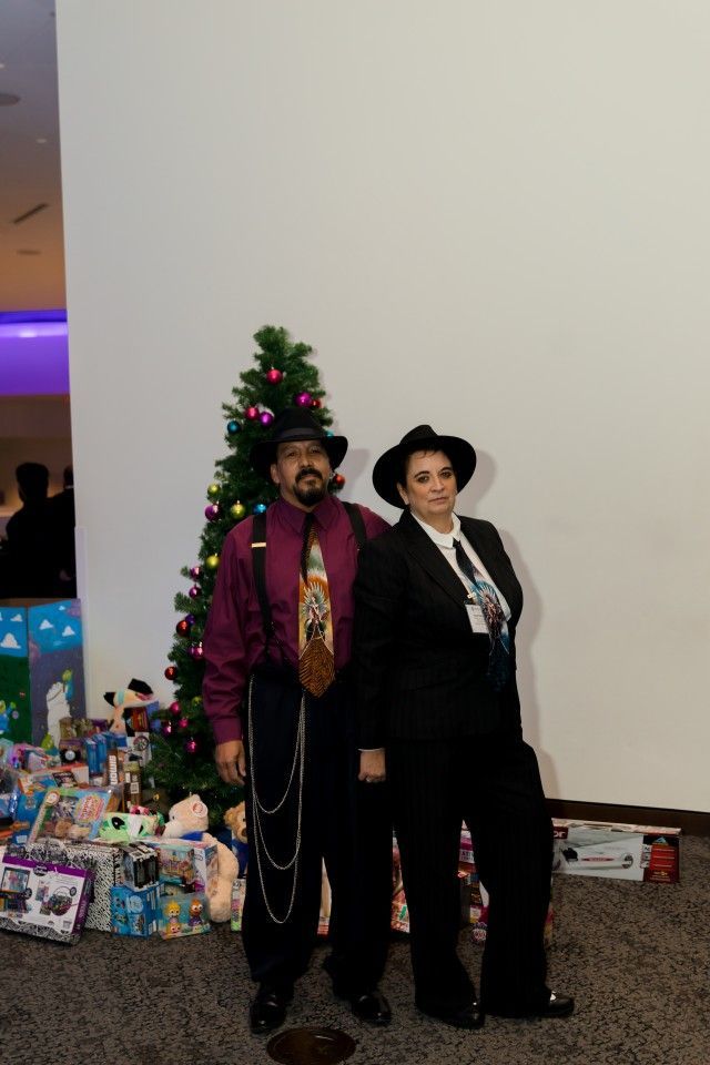 Two people in suits pose by a Christmas tree and gifts.