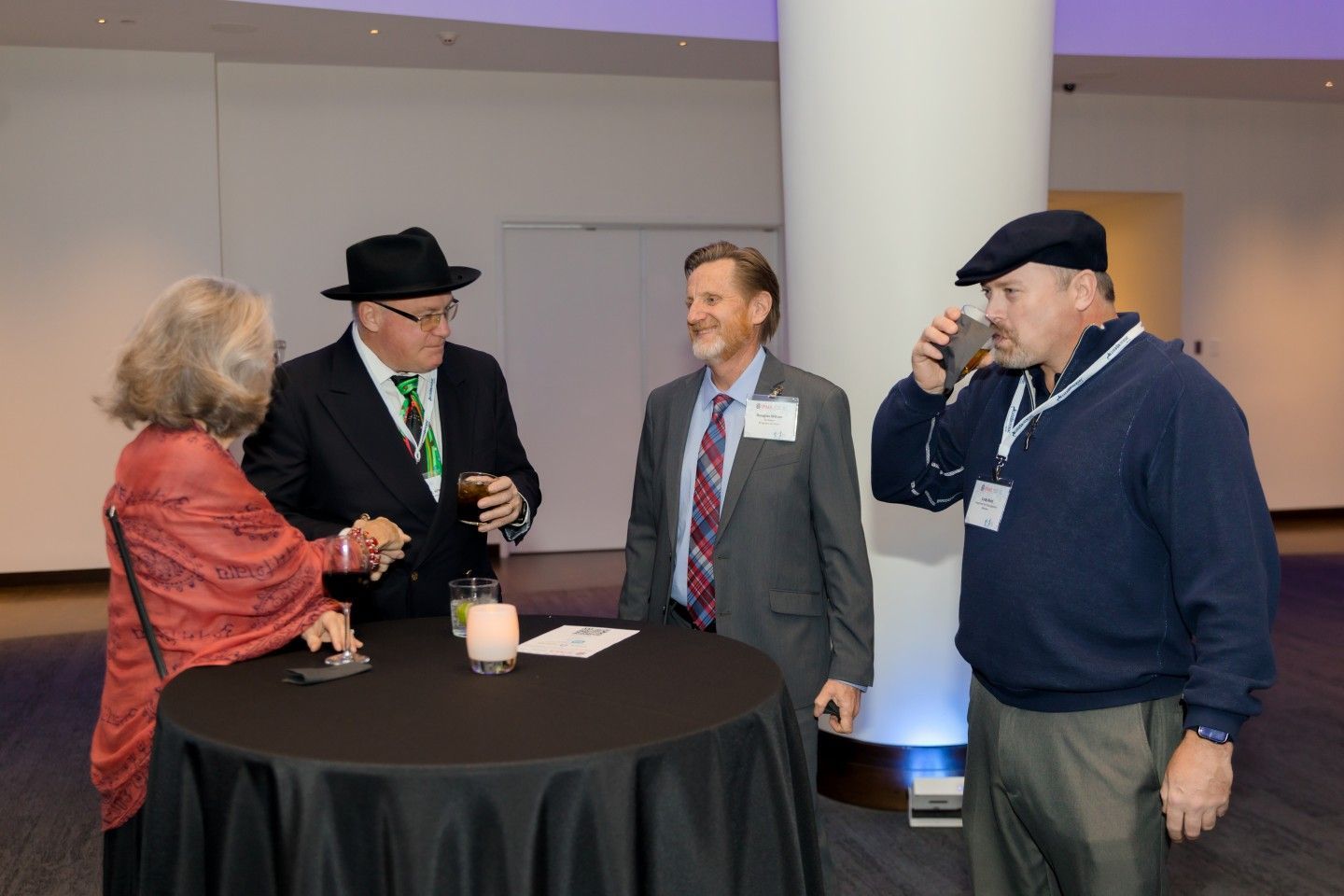 Four people at a cocktail table, conversing. One man drinks; another wears a hat, tie, and suit.