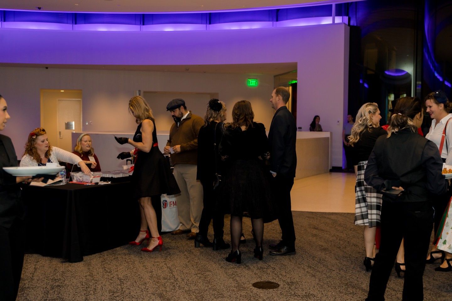 People gathering at a reception desk, some in formal attire. Venue has purple lighting and modern decor.