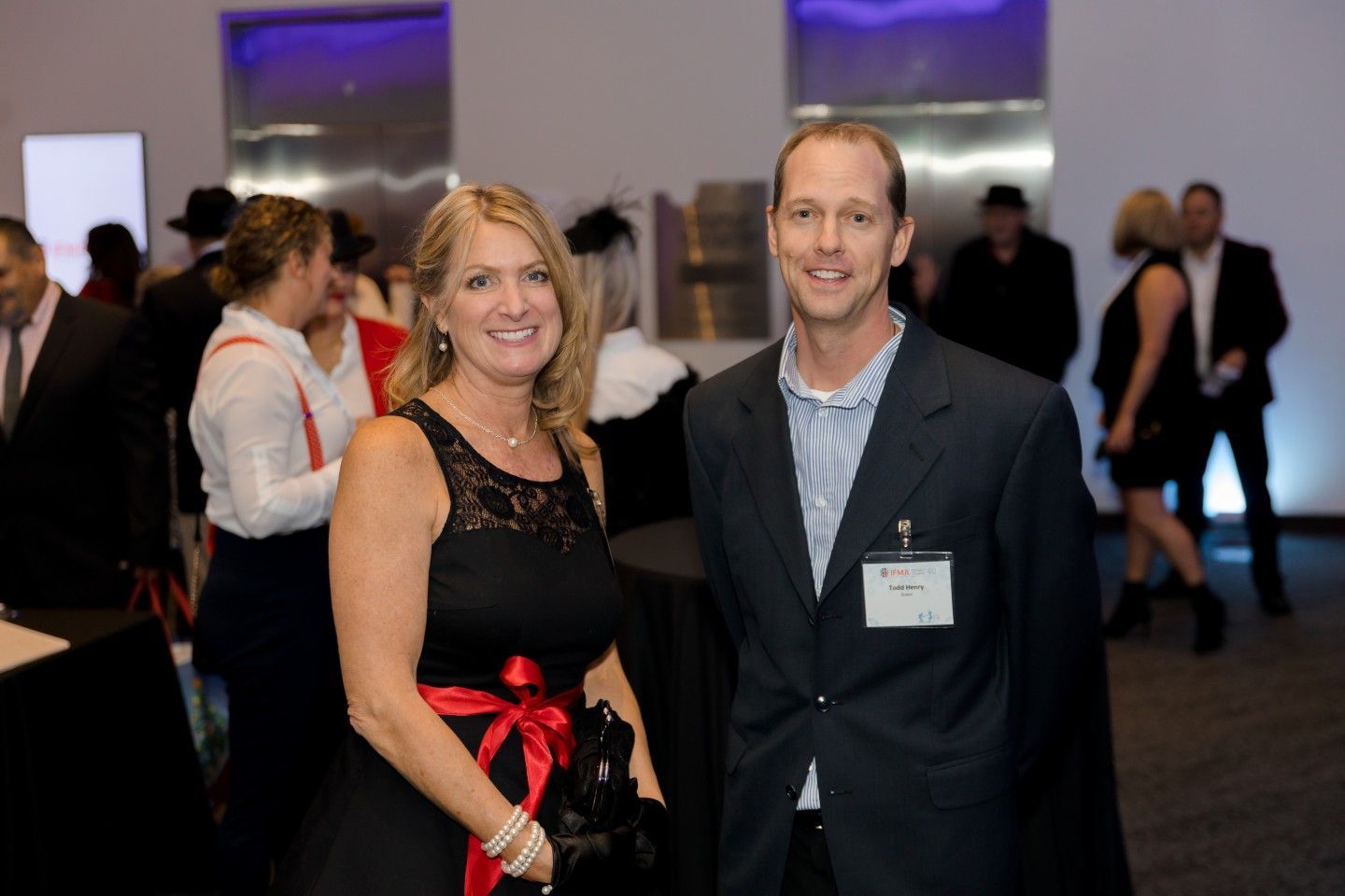 Woman in black dress and man in a suit jacket pose at a gathering. Others mingle in the background.
