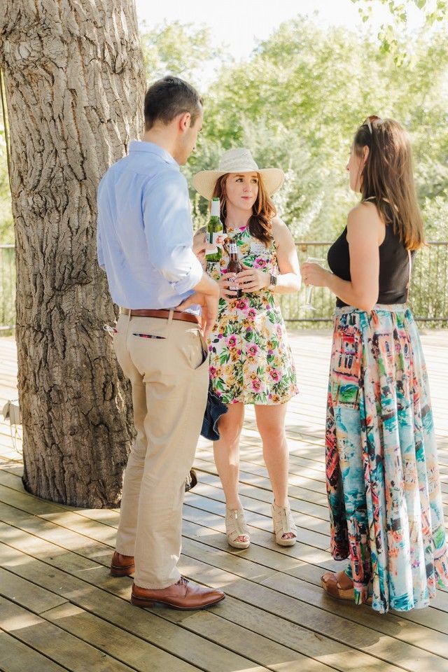 Three people on a wooden deck; a woman in a floral dress and hat talks to two others, outside.