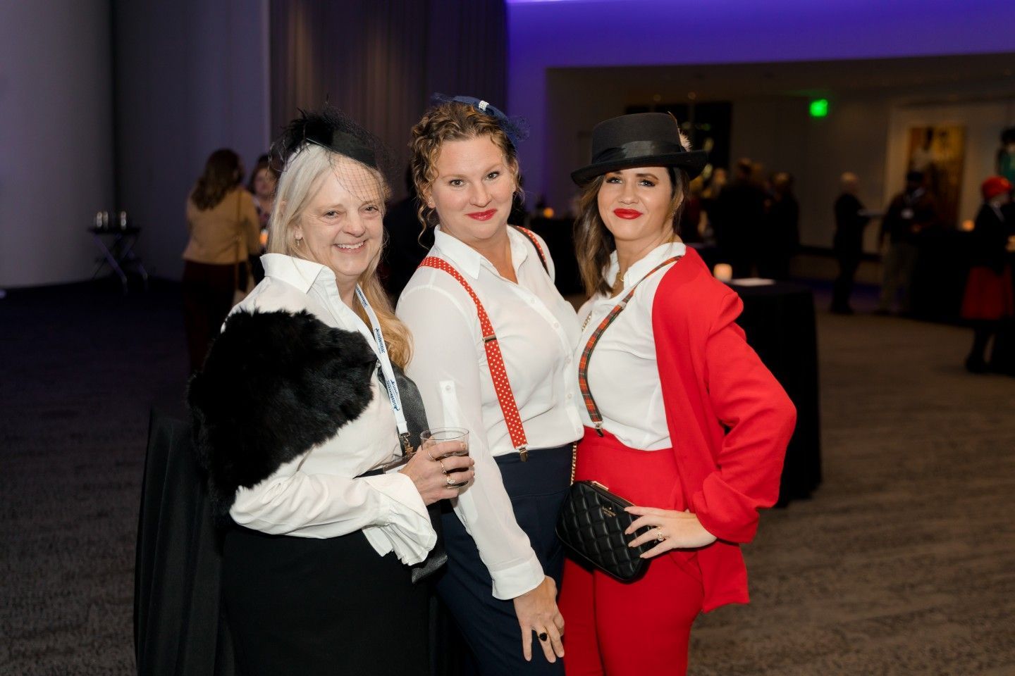 Three women in vintage-style outfits at an event. One wears a black hat, another suspenders, the third a red suit.