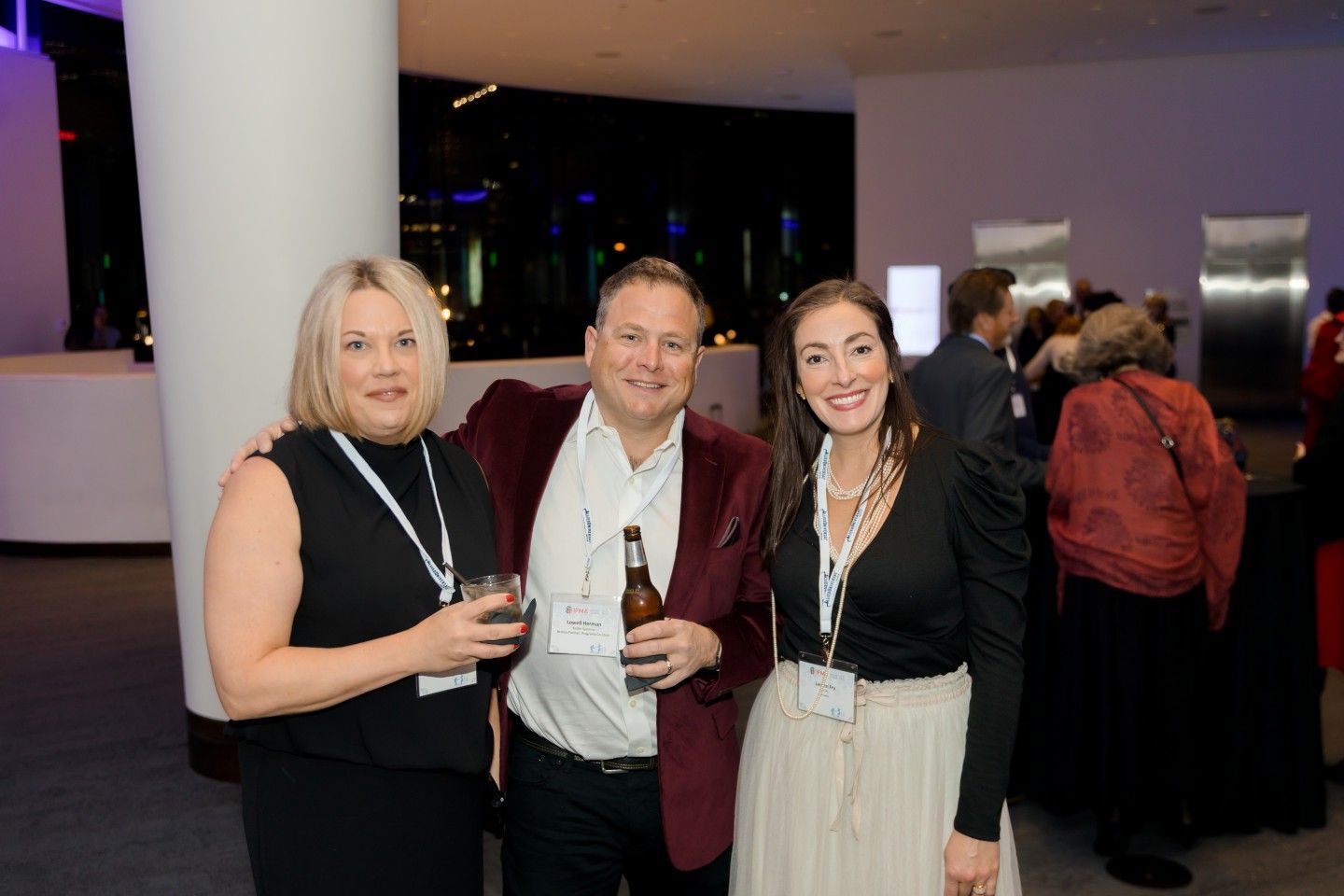 Three people at an event, posing and smiling; two women, one in black, the other with a white skirt; man in a maroon jacket.