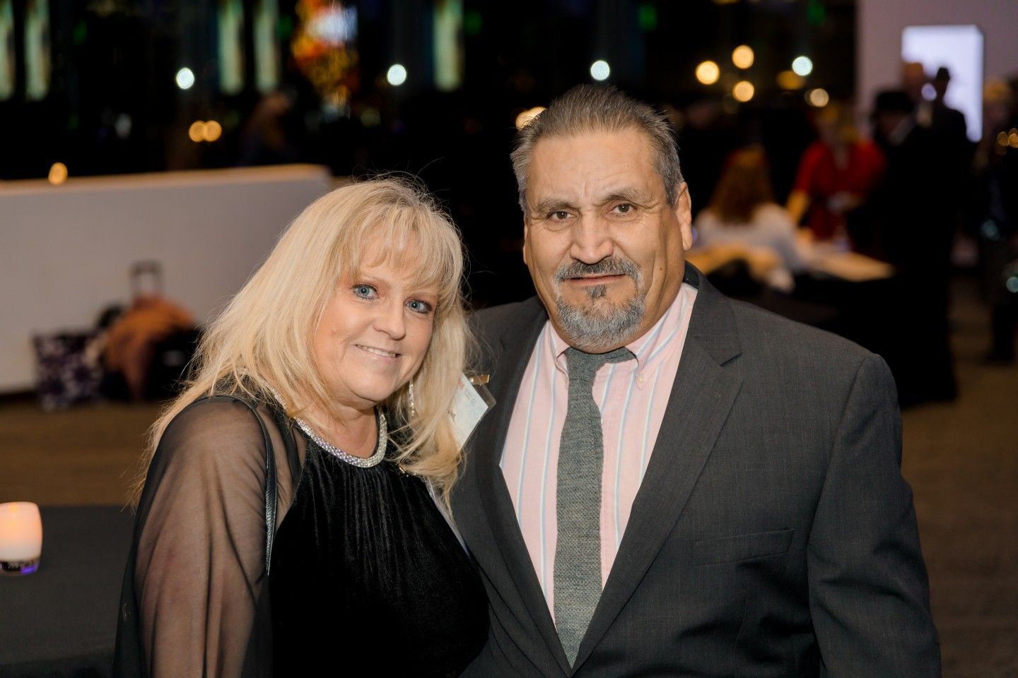 Couple smiling at a formal event, woman with blonde hair, man in suit. Dark setting.