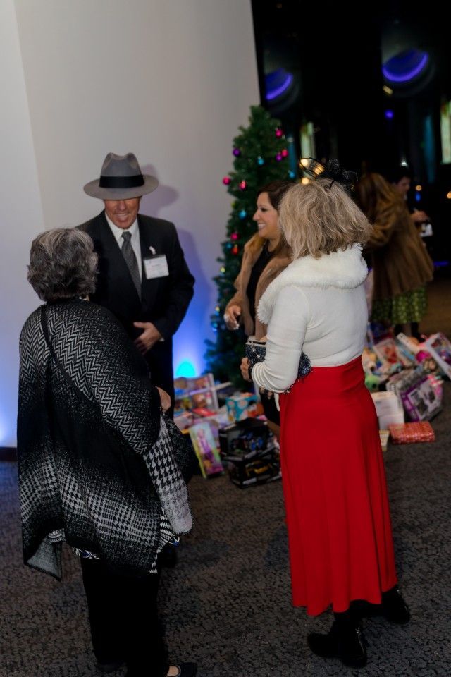 People at a gathering with gifts, Christmas tree; man in a hat; woman in a red skirt; indoor setting.