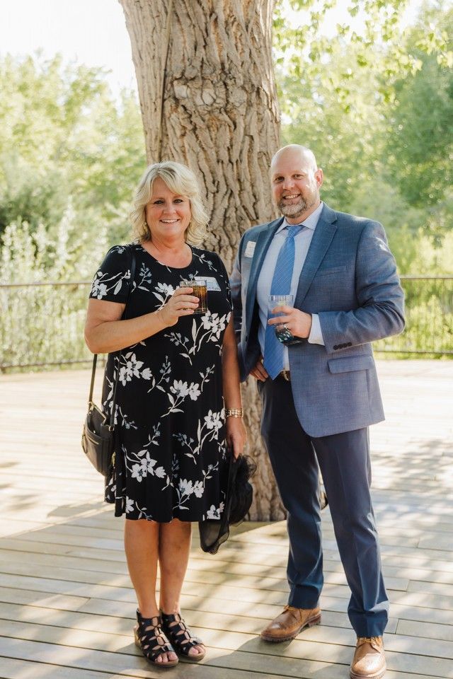 Woman in floral dress and man in a suit stand near a tree on a wooden deck, holding drinks.