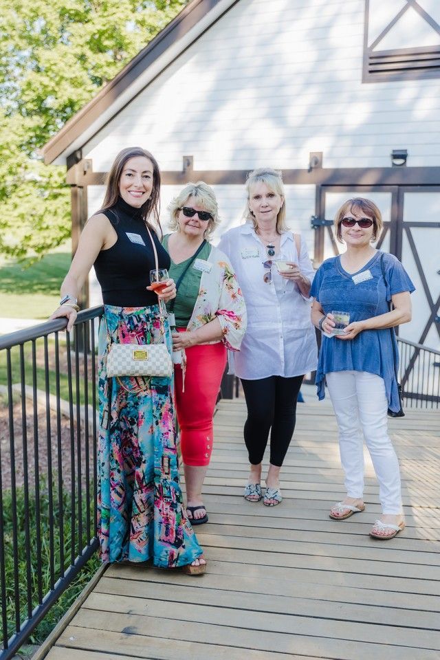 Four women smiling and posing on a wooden walkway. One holds a drink, building in the background.