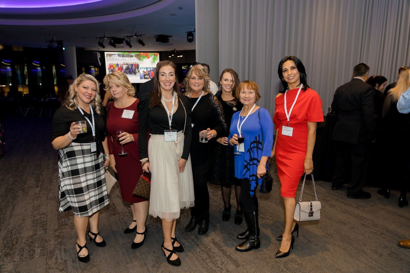 Group of women at an event, some holding drinks. Elegant dresses, varied colors, and a modern interior.