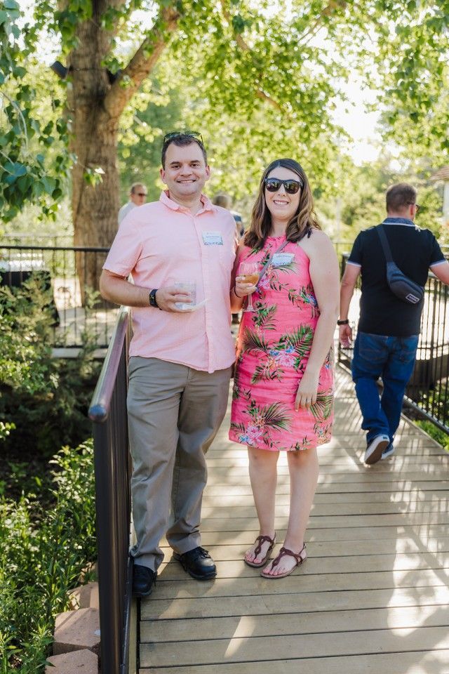 Couple standing on a wooden bridge, man in pink shirt, woman in floral dress, holding drinks, sunny outdoor setting.